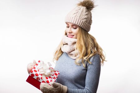 Beautiful young blonde woman in warm clothes with gift box on hand on white background. Happy girl wearing beige knitted hat, scarf, mittens and grey sweater. Christmas, new year, winter conceptの写真素材