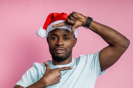 Closeup of calm serious unshaven afro american man making frame sign with both hands, wearing casual white t shirt, Santa hat, standing against pink background. Gesture concept.の写真素材
