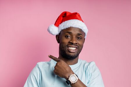 Cheerful unshaven african man wearing Santa hat, white t shirt, stylish wrist watch, pointing with index finger to side, showing something interesting, isolated on pink background. Advertising conceptの写真素材