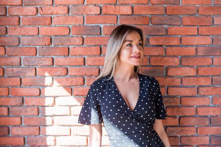 Happy cheerful young woman wearing in polka dots looking aside with joyful and charming smile near the red brick wallの写真素材