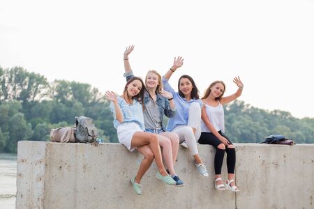 Happy female Friends Having Fun On Weekend, On Picnic Outdoors. Young Smiling People Sitting on concrete border and looking at cameraの写真素材