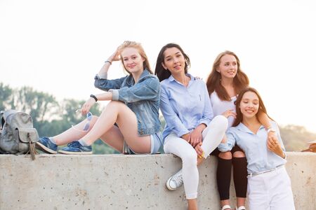 happy cute female friends sitting at concrete border and looking camera