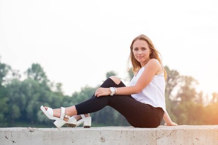 Portrait of beautiful young woman in white t-shirt and black trousers sitting in nature at sunset....