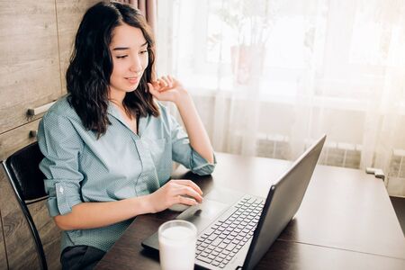 Portrait of young happy brunette female entrepreneur working online with black laptop while sitting at table in her home office dressed casually. Remote work, online education, technology concept.の写真素材