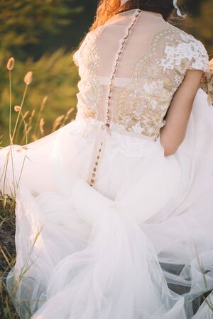 Closeup of a beautiful bride in a white handmade dress and with a decoration on her hair sitting on the grass in nature, from behindの写真素材