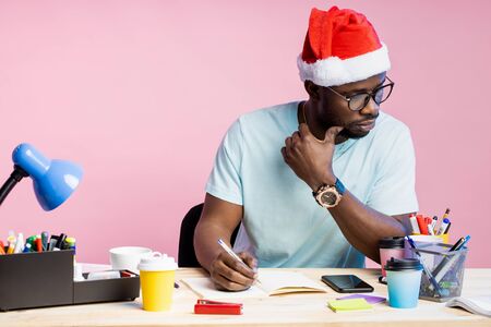 Serious young african student with small beard dressed in white t shirt, Santa Hat, sitting at table with office supplies, smartphone, doing homework, over pink background. Copy space.の写真素材