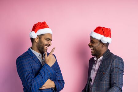 Joyful afro american entrepreneur and indian businessman smiling and laughing, standing over pink background, wearing Santa hats. Best friends having fun after work. Communication, friendship concept.の写真素材