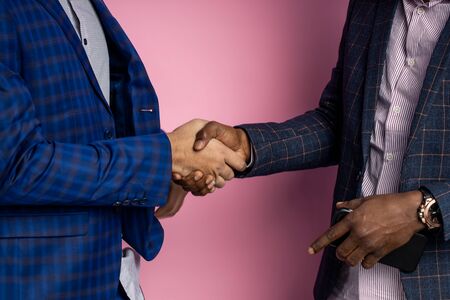 Closeup cropped shot of two multiethnic businesspeople wearing suits, shaking hands after signing good contract, congratulating each other, standing over pink background.の写真素材