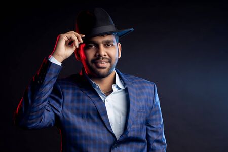 Studio portrait of young handsome confident unshaven Indian man, businessman wearing shirt, checked suit, looking at camera, touching hat, smiling, standing isolated on dark background.の写真素材