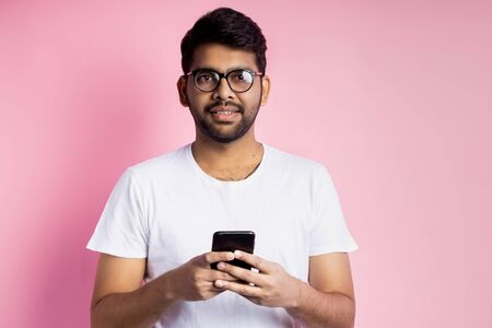 Portrait of good-looking happy indian young man in glasses, holding cell phone, sending text message, reading good news, posing against pink background. Technology, communication.の写真素材