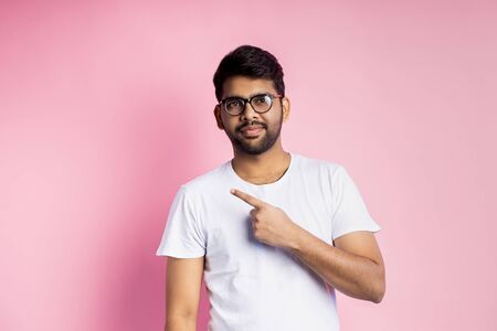Attractive unshaven young indian man wearing white t shirt, eyewear, pointing with index finger to side, smiling, showing something interesting, isolated on pink background. Advertising concept.の写真素材