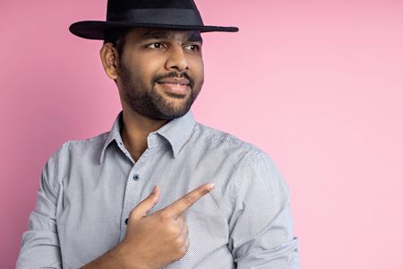 Close up shot of pleased joyful young indian male with stubble, wearing casual shirt, black hat, looking with smile and pointing to side with forefinger, standing against pink studio wall.の写真素材