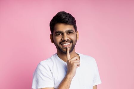 Horizontal close up portrait of handsome unshaven young indian man, student wearing white t shirt, looking at camera with broadly smile, keeping index finger on chin, isolated over pink background.の写真素材