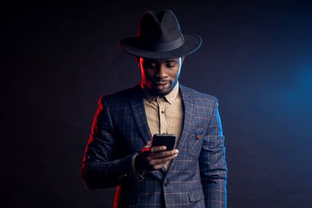 Waist up shot of confident stylish african guy, holding mobile phone, looking at screen, reading...