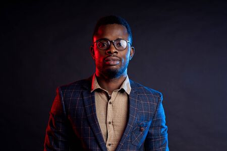 Closeup portrait of young handsome confident serious bearded african businessman in glasses, wearing shirt, checked suit, looking at camera, standing against black background.の写真素材