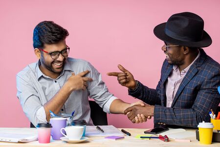Happy african american and indian business partners sitting at desk in office and shaking hands,...