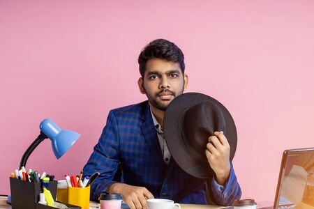 Indian businessman wearing suit, black hat, looking at screen of mobile phone, waiting for important call, reading news, sending email, sitting at working desk in office, over pink background.の写真素材