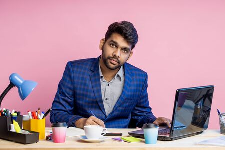 Serious confident young manager sitting at desk surrounded with stationery, dressed in suit, using laptop, writing something in notepad, making report, posing on pink background. Business concept.の写真素材