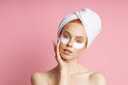 Portrait of beautiful young woman with smooth soft skin applying white under eye hydro gel patches. Charming lady with eye mask, towel on head, keeping palm on cheek posing against pink studio wall.の写真素材
