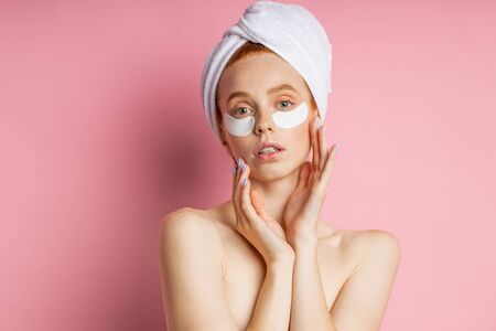 Headshot of sensual beautiful woman applying patches to reduce under eye puffiness and dark circles, touching gently face with both palms, posing naked isolated on pink background. Beauty concept.の写真素材