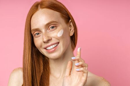 Portrait of cheerful beauty model has fresh pure skin, applying wrinkle cream, smiling broadly with white teeth, looking with happy expression on pink background. Beauty product, skin care, wellness.の写真素材