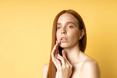 Closeup of tender relaxed caucasian young woman with red hair, bare shoulders, freckled clean skin touching cheek with hand isolated on yellow background, looking at camera. Skin care, beauty concept.の写真素材