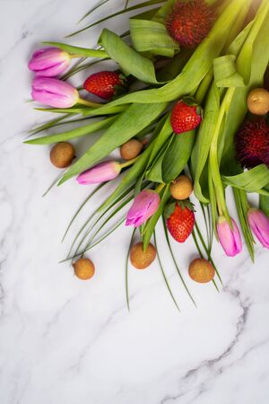 Top view beautiful pink tulips with small exotic fruits on a marble background on a table with a copy space. Longan, rambutan, strawberry with flowersの写真素材