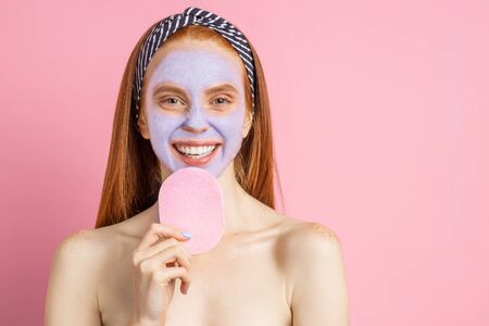 Horizontal shot of happy overjoyed woman standing naked, with mask for rejuvenation on face, having cosmetic treatments, smiling showing white teeth, keeping sponge, posing against pink studio wall.の写真素材