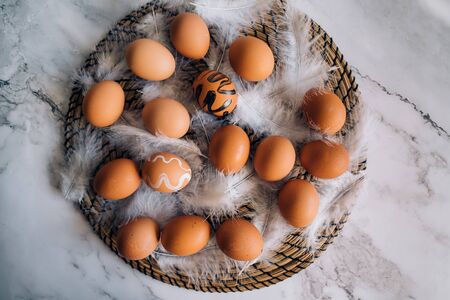 Happy Easter background. Top view composition of brown chicken eggs and white fluffy birds feathers on round wicker stand on marble table. Easter holiday decorations, flat lay.の写真素材