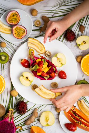 Top view of plates with sliced fruits. Assortment of different exotic fruits on marble table. Delicious breakfast, healthy food, summer. Female hand holding ripe strawberry. Flat lay.の写真素材
