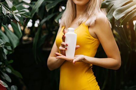 Gorgeous caucasian blonde woman in yellow swimsuit holding cosmetic product, bottle of sunscreen lotion, standing among tropical plants. Beauty, cosmetics, advertising concept. Cropped photoの写真素材