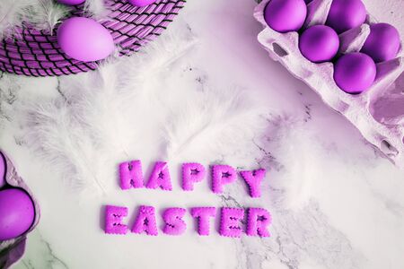 Creative bright composition with festive atmosphere. Easter holiday decorations on marble background. Fluffy white feathers and painted purple eggs in carton box and on table.の写真素材