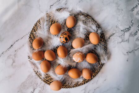 Happy Easter background. Top view composition of brown chicken eggs and white fluffy birds feathers on round wicker stand on marble table. Easter holiday decorations, flat lay.の写真素材