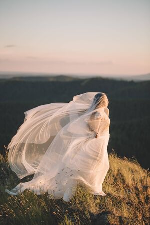Portrait of a beautiful young brunette bride woman in a white dress and with a long veil Curling in the wind in nature, the mountains in summer. Fine art, wedding, lifestyle, love, life conceptの写真素材