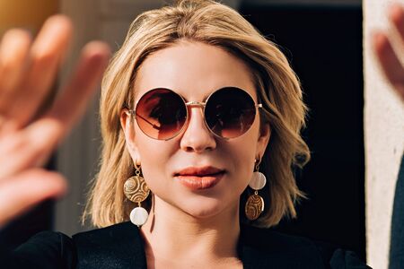 Lovely caucasian woman with short haircut in stylish sunglasses, day makeup, taking a selfie walking around town while traveling. Outdoor closeup photo of cute amazing girl enjoying spring.の写真素材