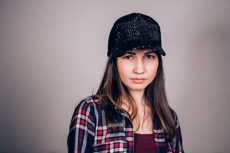 Portrait of a beautiful young girl in a shirt and a black baseball cap with sequins on a gray background. The girl looks at the camera with a serious lookの写真素材