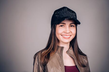 Beautiful young brunette woman with a charming smile looking at the camera. Girl wearing a black baseball cap and brown leather jacket. Positive emotionsの写真素材