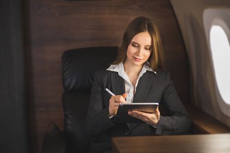 Attractive caucasian businesswoman sitting in comfortable seat in airplane, holding modern tablet, working using wireless connection on board. Business, traveling and technology.の写真素材