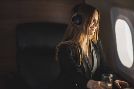 Happy cheerful sexy woman with long blonde hair, in a stylish suit, sitting in an airplane seat, looking out the window, listening to music with headphones, enjoying the flight.の写真素材