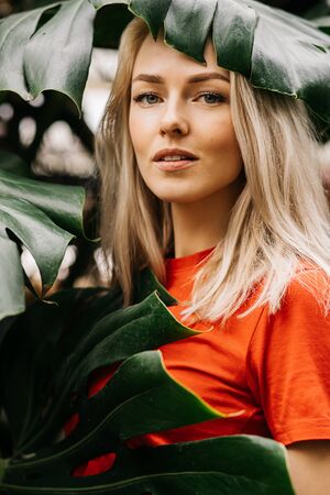 Blonde lady with natural makeup posing in rainforest. Gorgeous caucasian woman in red t shirt standing among monstera leaves, enjoying summer vacation. Wildlife and people concept.の写真素材