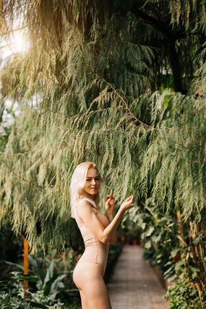 Beautiful smiling slender woman in flesh color swimsuit standing under leaves of big exotic tree. Slim blonde lady happy to spending vacation at resort in rainforest. Body care.の写真素材