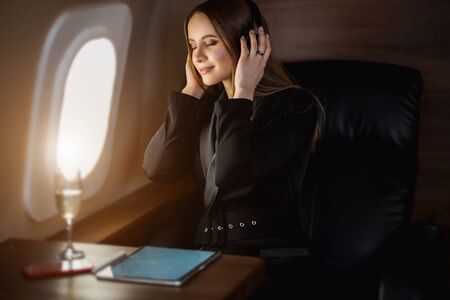 Happy caucasian young blonde woman in stylish black dress relaxing during flight in private jet,...
