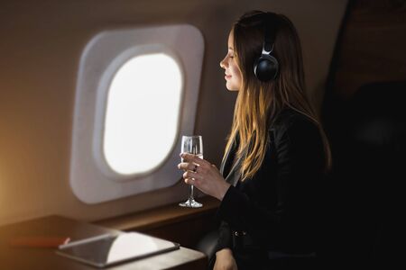 Closeup shot of happy young caucasian woman with long blonde hair, in black jacket dress, listening to music with headphones, holding glass of champagne, looking at airplane window with cute smile.の写真素材