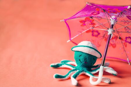 Cute crochet octopus doll on a coral background. Playful knitted green octopus wearing white hat sitting under umbrella. Handmade knitted children toy. Summer, vacation concept.の写真素材