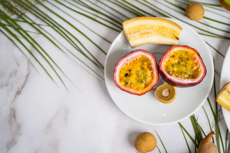 Sliced passion fruit, longan, banana on white dish. Exotic fruits and tropical leaves on white marble background with copy space for your text. Food, summer concept. Top view.の写真素材