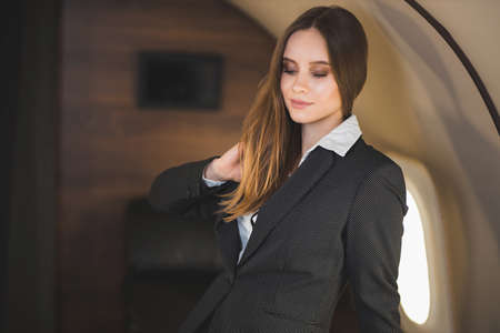 Beautiful caucasian young brown haired lady with nice makeup, wearing stylish black suit, looks down, touches her hair and gently smiles, poses in private aircraft. Traveling at first class.の写真素材