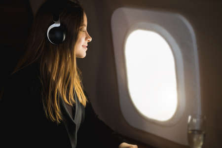 Attractive happy young woman with long blonde hair, in black jacket dress, sitting in an airplane seat with closed eyes, listening to music with headphones, enjoying the flight.の写真素材