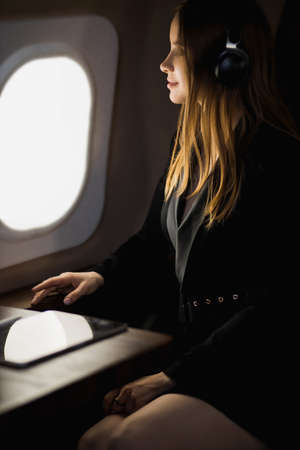 Happy caucasian young blonde woman in stylish black dress relaxing during flight in private jet, listening music using wireless headphones with closed eyes.の写真素材