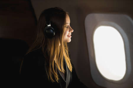 Attractive happy young woman with long blonde hair, in black jacket dress, sitting in an airplane seat with closed eyes, listening to music with headphones, enjoying the flight. Focus in headphonesの写真素材