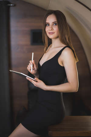 Lovely slender young woman wearing sexy black dress in private jet. Successful young woman entrepreneur holding digital tablet, working during the flight. Technology concept.の写真素材